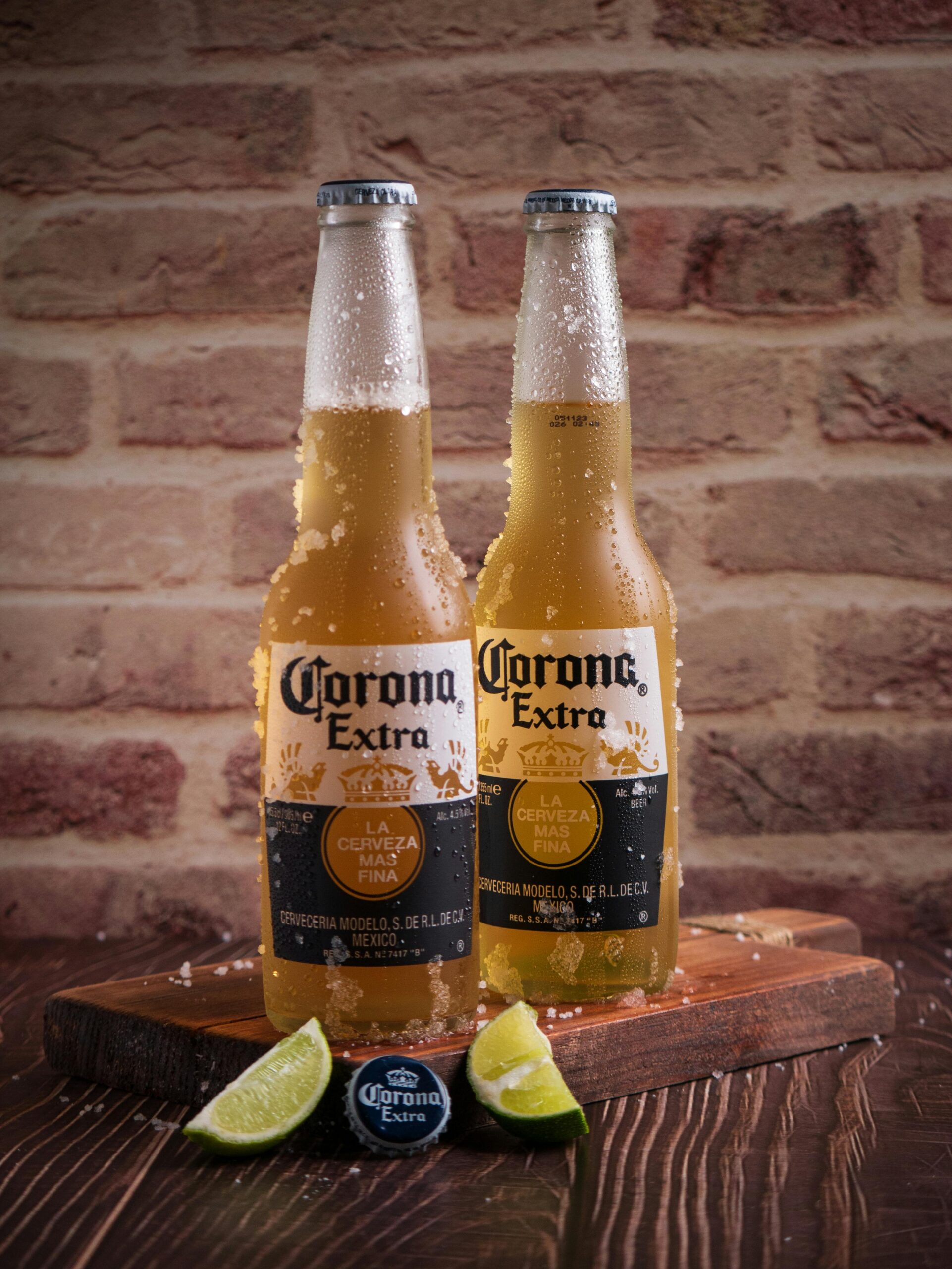 Cold Corona Extra beer bottles on a wooden table with lime slices against a brick wall backdrop.